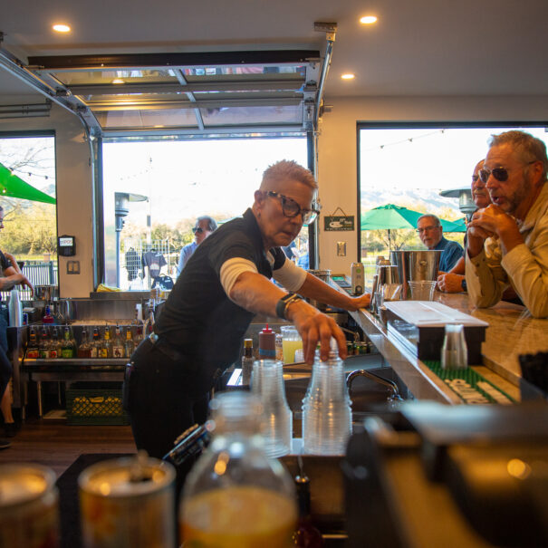 A bartender serves drinks to customers seated at a bar counter. Other patrons and staff are visible in the background. The setting appears to be a well-lit, casual bar with an outdoor view.