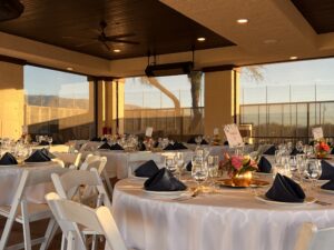 A beautifully decorated outdoor event space with round tables covered in white linens and navy blue napkins. The space has a vaulted ceiling with exposed wood beams and large windows overlooking a scenic view. There are hanging lights and ceiling fans, creating a cozy and inviting atmosphere.