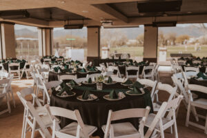 The pavilion at forty niner country club, outdoor event space with round tables covered in black linens and white chairs. The space has a covered patio with large windows overlooking a golf course. Green napkins and small floral centerpieces adorn the tables.