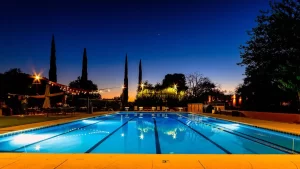 The pool at forty niner country club illuminated by lights at night. The pool is surrounded by trees and a building with lights visible in the background.